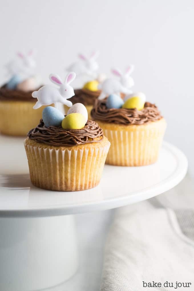 Four Easter Egg Cupcakes (yellow cake cupcakes topped with chocolate buttercream frosting, 3 Cadbury mini eggs, and an Easter bunny party pick) arranged in a semi-circle on a white cake stand