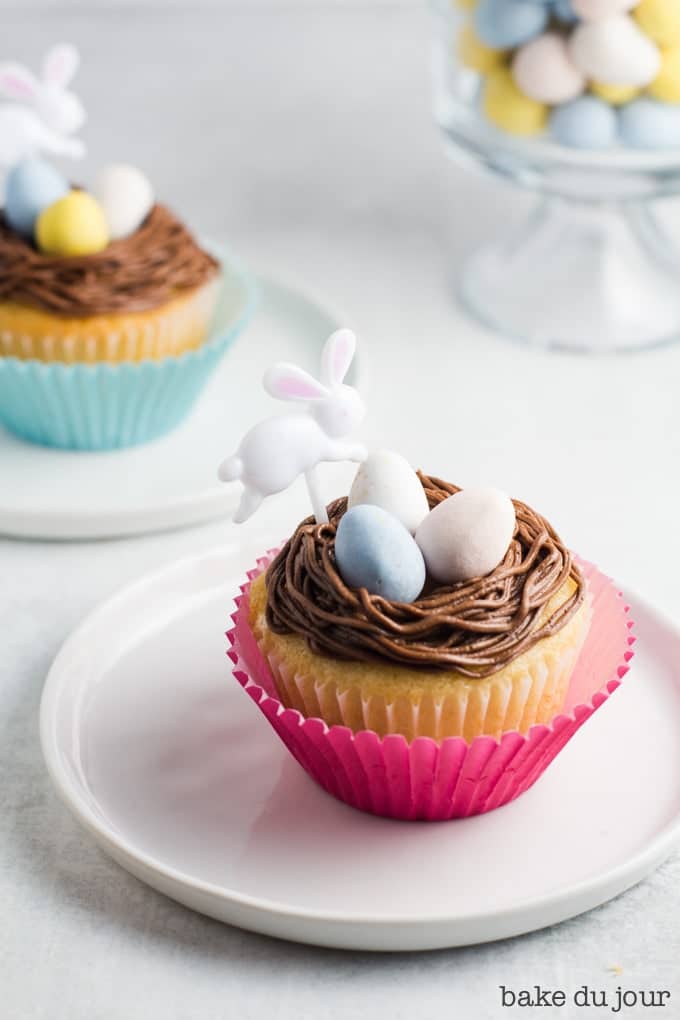 A yellow cake cupcake topped with chocolate buttercream frosting, three Cadbury mini eggs, and an Easter bunny party pick, with another cupcake in the background