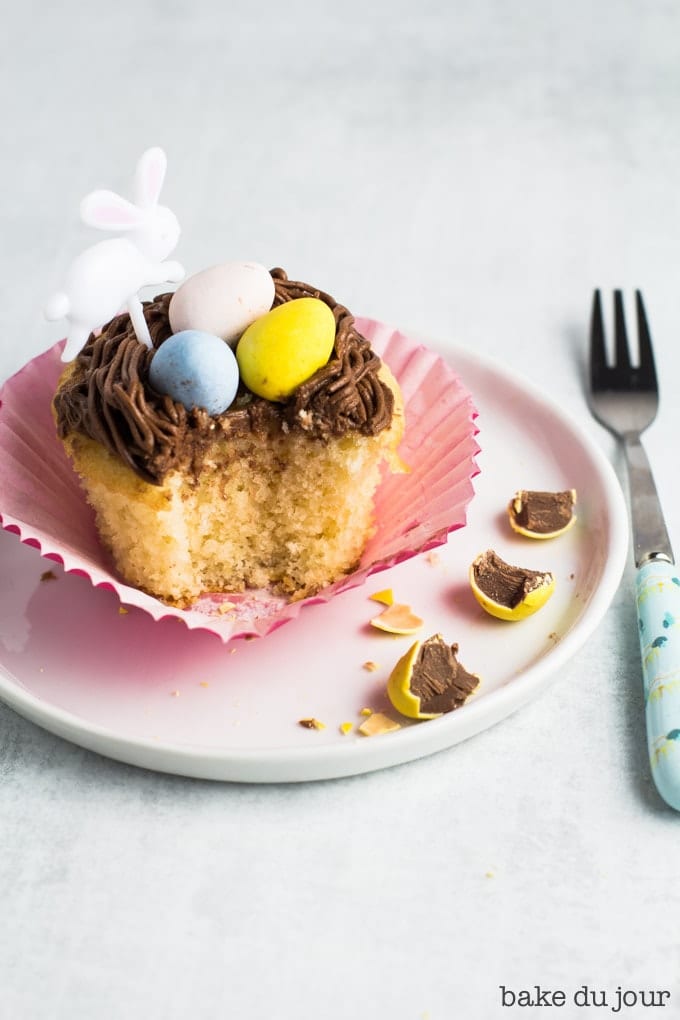 A yellow cake cupcake topped with chocolate buttercream frosting and an Easter bunny party pick on a white dessert plate, with a bite taken out of it. A light blue dessert fork placed on the right of the plate